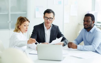 Drei Personen sitzen an einem Tisch und betrachten Dokumente und einen Laptop.