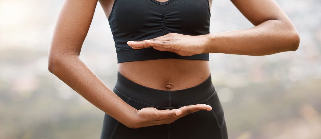 Person hält Hände vor dem Bauch. Schwarzes Sport-Outfit. Fokus auf den Bauchbereich. Hintergrund unscharf.