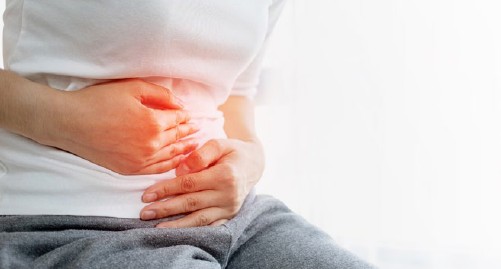 Person sitzt, Hände auf dem Bauch. Roter Bereich auf dem Bauch. Graue Hose, weißes Oberteil.