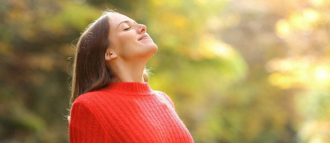 Eine Frau atmet tief ein. Sie steht im Freien, im Hintergrund Bäume. Sie trägt einen roten Pullover.