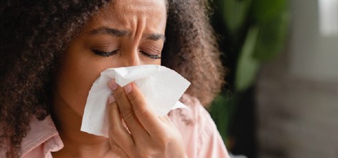 Eine Frau putzt sich die Nase mit einem Papiertaschentuch. Sie hat die Augen geschlossen und runzelt die Stirn.
