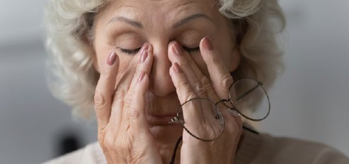 Eine ältere Frau reibt sich die Augen. Sie trägt eine Brille und scheint müde zu sein.