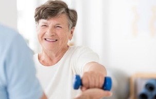 Ältere Frau trainiert mit einer blauen Hantel. Sie lächelt. Eine weitere Person ist zu sehen.