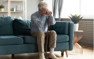 Ein älterer Mann sitzt auf einem Sofa und stützt sich auf einen Stock. Er wirkt traurig.