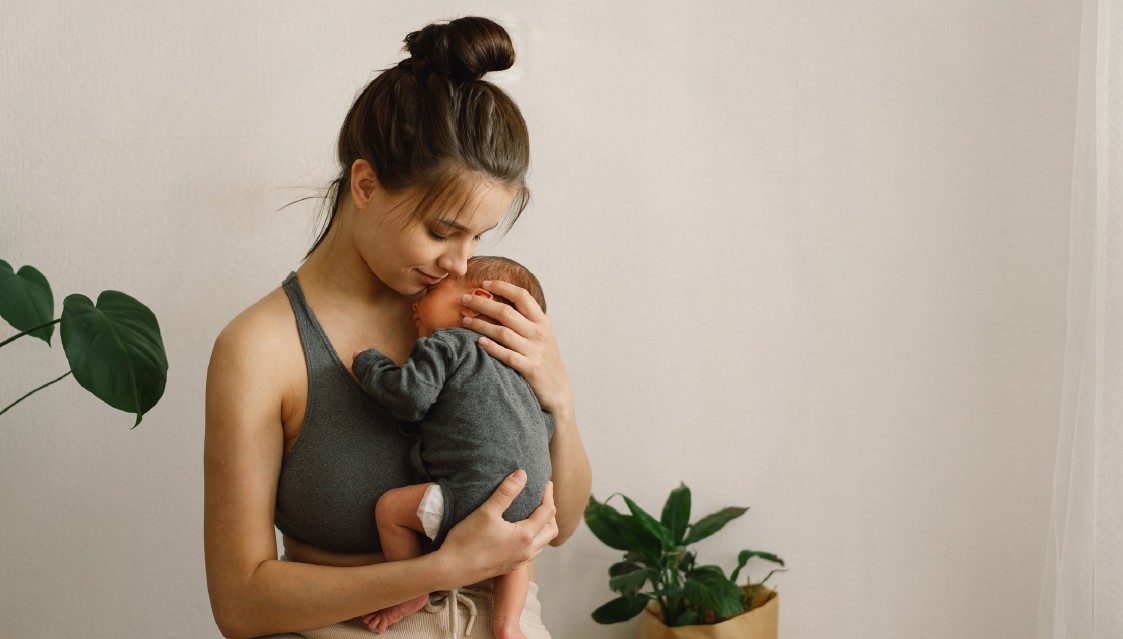 Frau hält Baby im Arm. Sie trägt ein graues Top. Baby in grauem Strampler. Weißer Hintergrund.