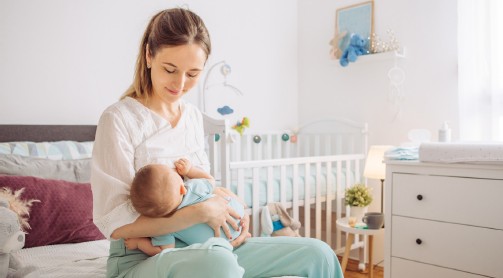 Frau stillt Baby im Bett. Baby in blauem Strampler. Weißes Zimmer mit Spielzeug und Kommode.