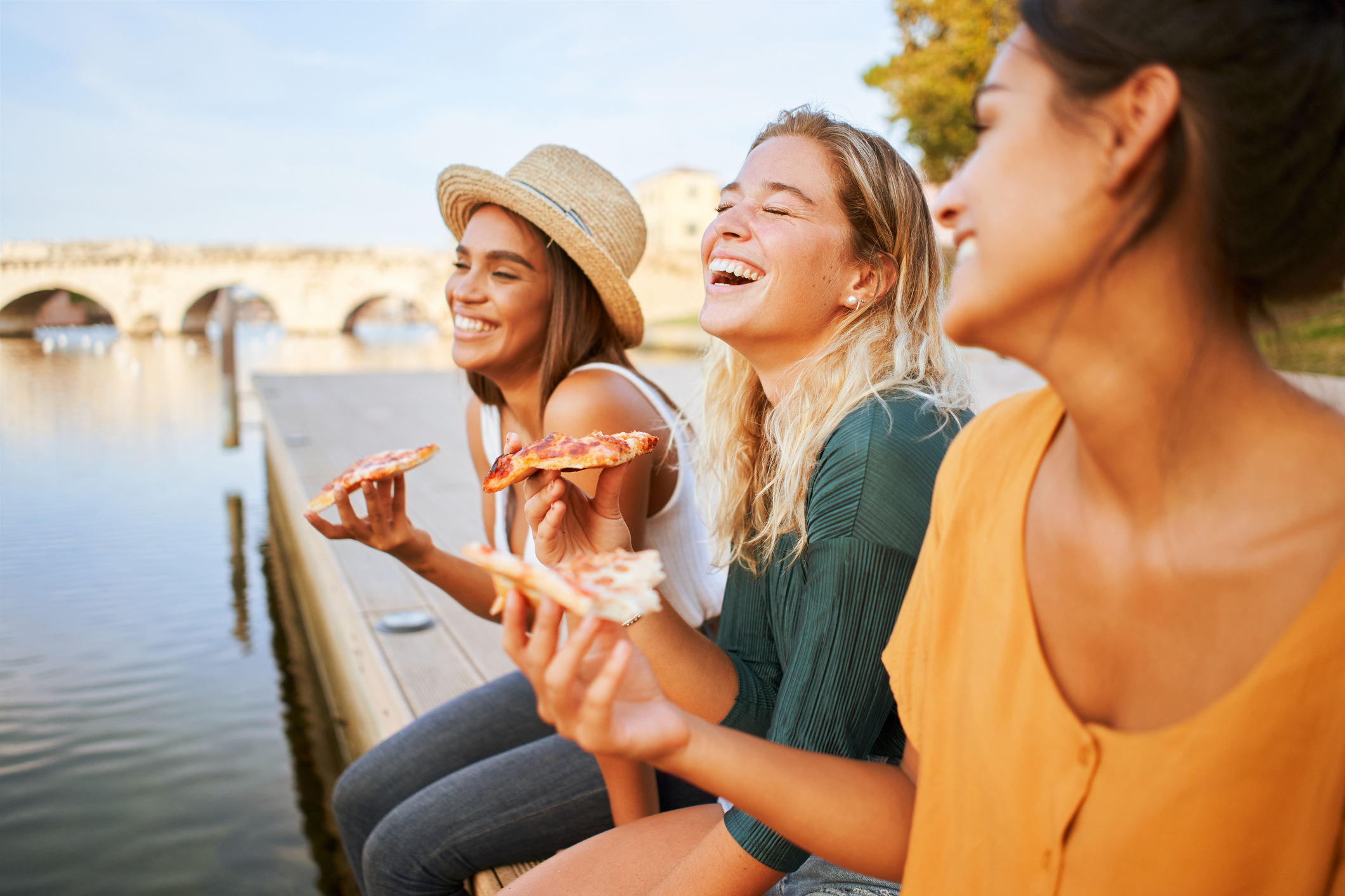 Drei junge Frauen sitzen am Wasser und essen Pizza. Sie lachen und unterhalten sich.