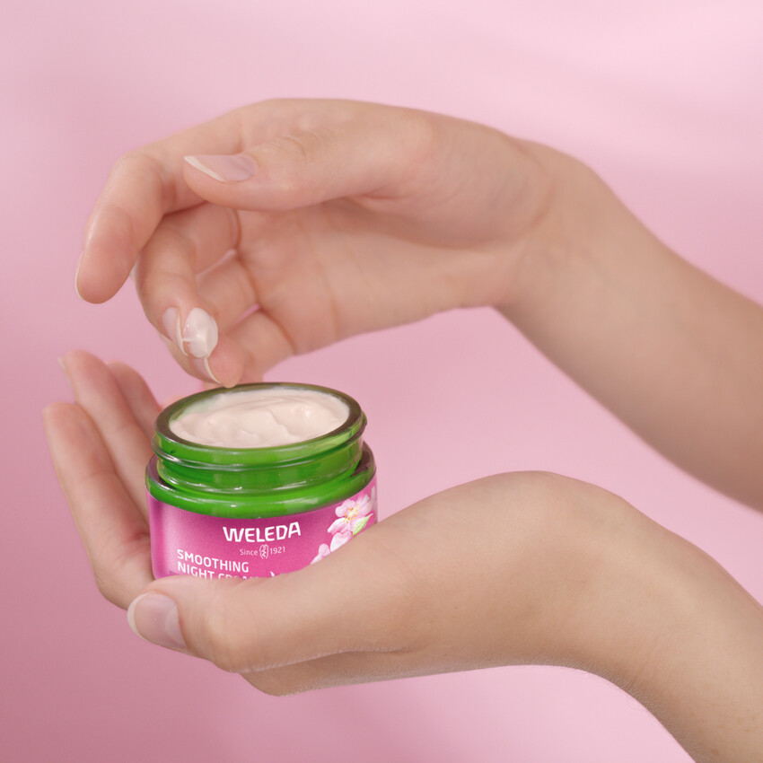 Hände halten Creme-Tiegel. Weleda Smoothing Night Cream. Rosa Etikett, grüner Deckel. Creme wird entnommen.