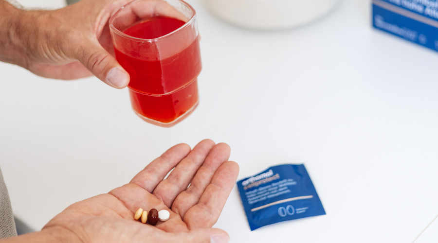 Person hält Glas mit roter Flüssigkeit und Tabletten. Weißer Tisch, helle Umgebung.
