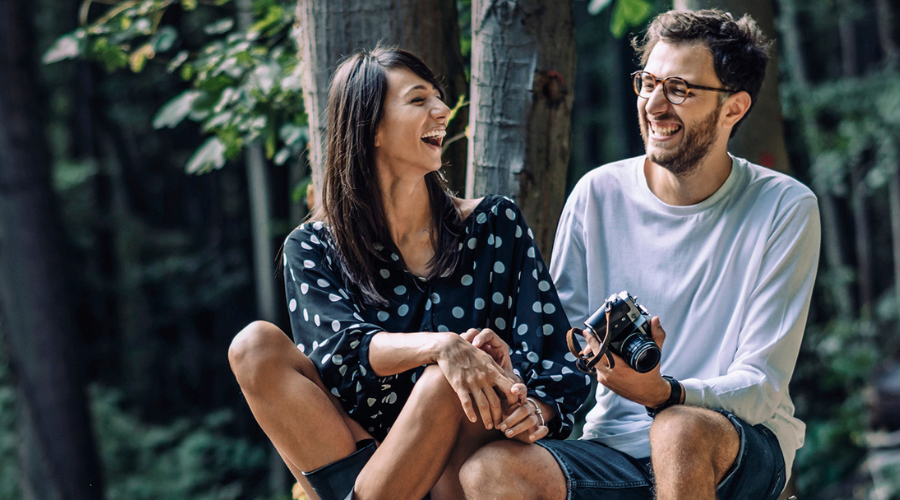 Paar lacht. Mann mit Brille und Kamera. Frau mit dunklem Kleid. Waldhintergrund.