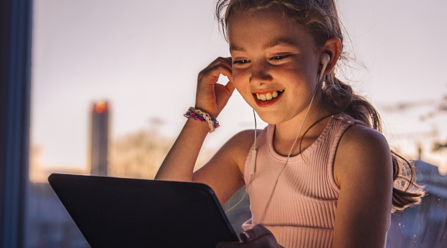 Mädchen mit Tablet, lächelt. Trägt Kopfhörer. Hintergrund: Fenster mit Sonnenlicht.