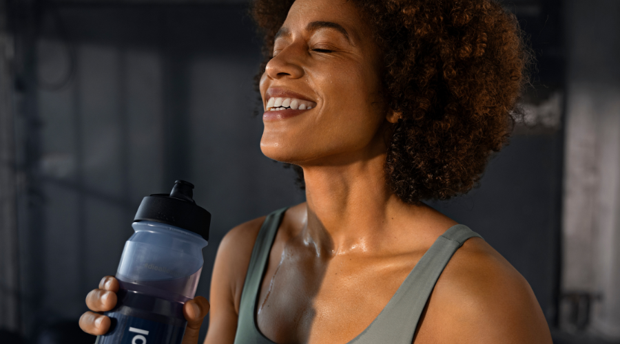 Frau trinkt aus einer Flasche. Schweiß auf der Haut. Orthomol Sport perform.