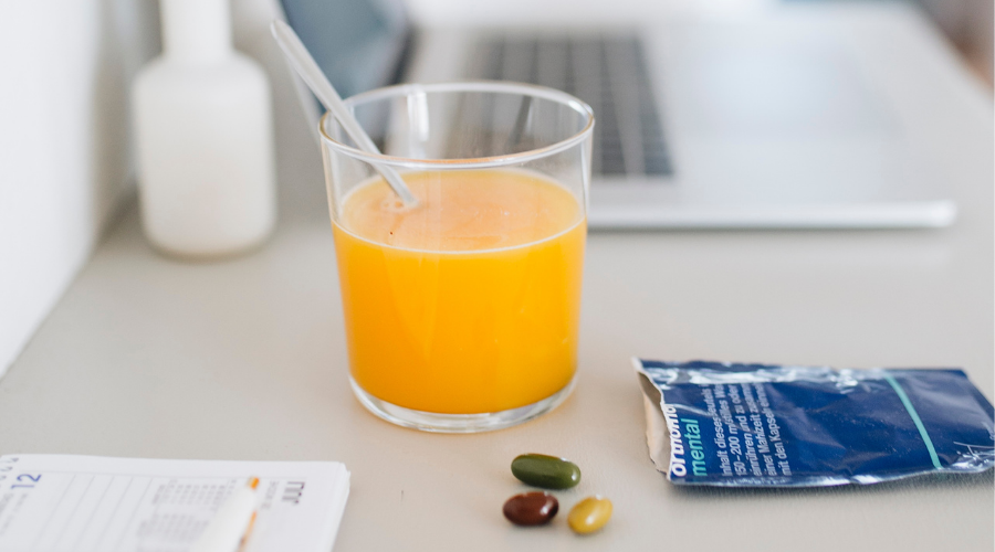 Glas Orangensaft, Kapseln und Beutel. Auf dem Tisch: Laptop, Notizblock, Stift.