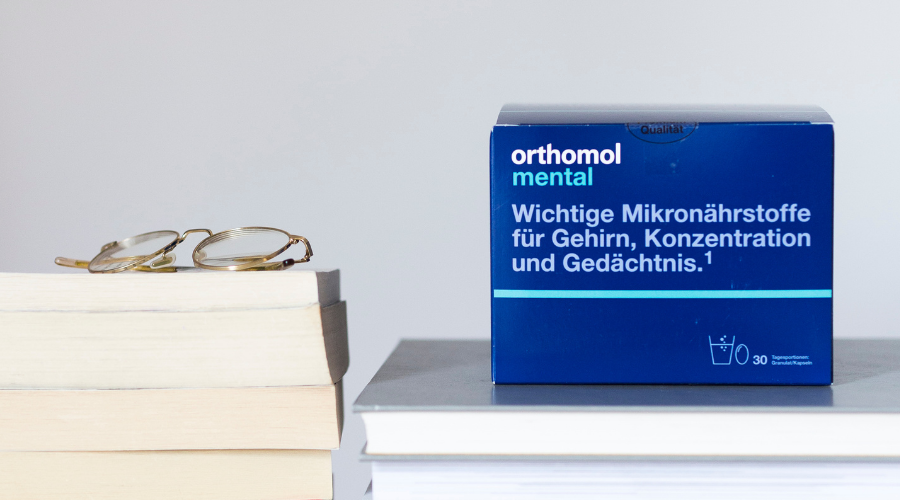 Orthomol mental-Packung, Brille, Bücherstapel. Text: Wichtige Mikronährstoffe für Gehirn, Konzentration und Gedächtnis.