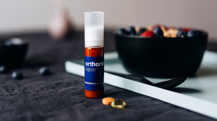 Orthomol Sport Flasche, Tabletten und Kapseln. Auf einem Tisch mit Beeren und Löffel.